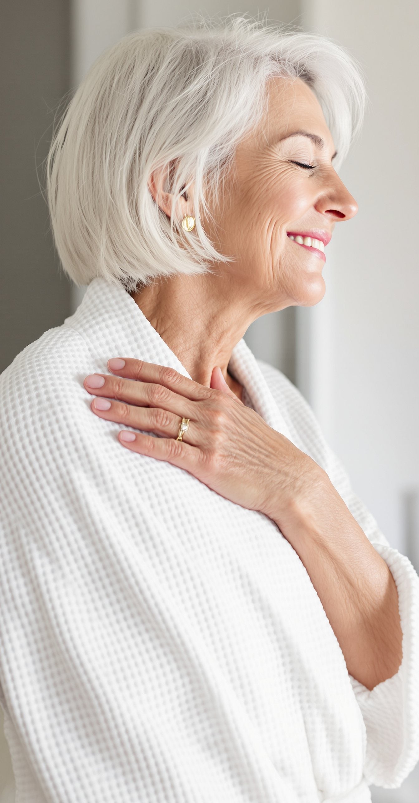 Woman with silver hair smiling with her eyes closed, resting one hand on her shoulder and neck